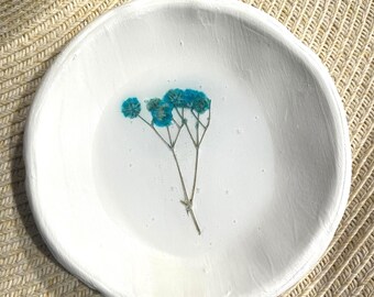 May include: A white, round, handmade ceramic dish with a textured surface. The dish contains a sprig of dried blue flowers encased in clear resin. The dish is set on a woven straw surface.