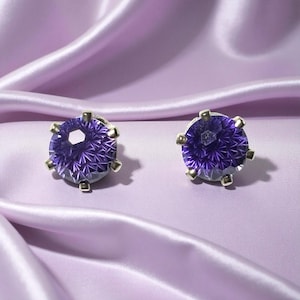 May include: A pair of stud earrings featuring large, faceted, round, purple gemstones. The gemstones are set in a gold-colored metal with six prongs. The earrings are displayed on a lavender satin fabric.
