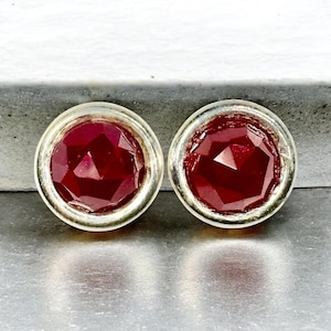 May include: A pair of stud earrings featuring faceted, deep red gemstones set in a silver-colored metal. The gemstones have a geometric cut, reflecting light. The earrings are round and have a simple, elegant design.