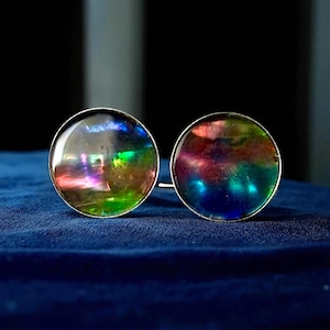 May include: A pair of round, iridescent cufflinks with a silver-colored border. The cufflinks display a spectrum of colors, including green, blue, purple, and red, against a dark blue background. The cufflinks are likely made of glass or a similar material.