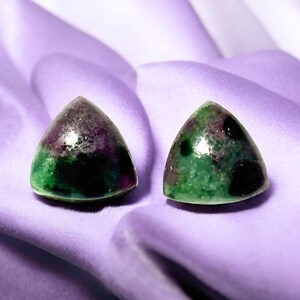 May include: Two triangular green, black, and purple gemstones on a purple fabric background.
