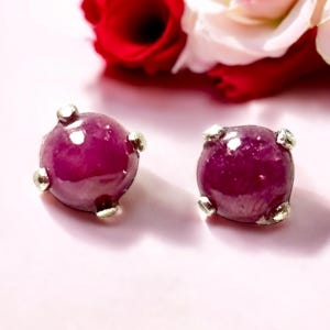 May include: A pair of stud earrings featuring round, cabochon-cut deep magenta gemstones. Each stone is secured by four small, silver-toned metal prongs that hold the gem in place. The surface of the stones appears smooth and polished.