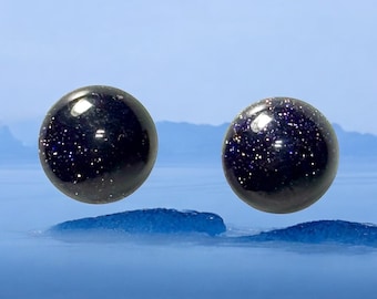 May include: Two dark blue, round gemstone beads with sparkling inclusions, possibly aventurine or a similar material. The beads are set against a backdrop of a blue sky and water, creating a serene and natural aesthetic.