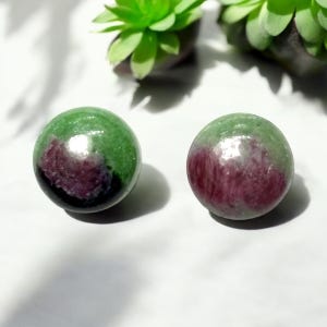 May include: Two polished gemstone spheres. Each sphere is a mix of green and burgundy colors. The spheres are smooth and reflective, with a glossy finish. The background includes green plants.