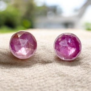 May include: Two pink gemstone earrings with a faceted cut. The earrings are round and have a shiny finish.