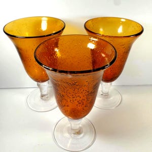 May include: Three amber-colored stemmed glasses with a bubble texture. The glasses have a flared bowl shape and a clear stem and base. The rims of the glasses are outlined with a dark brown color.