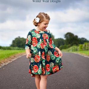 May include: A young girl wearing a dark blue dress with a floral print of red, green, and white roses. The dress has a pleated bodice and 3/4 sleeves. The girl is standing on a paved road with green grass and trees in the background. The text "Squared Away Tunic and Dress Size 1-16 Winter Wear Designs" is at the top of the image.