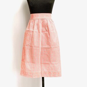 Puede incluir: Un delantal de vichy rosa y blanco con cintura fruncida y dos bolsillos. El delantal se muestra en un maniquí negro con un collar de perlas.