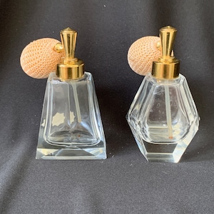 May include: Two clear glass perfume bottles with gold tops and a textured beige spray nozzle. The bottles are on a black background.