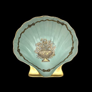 Op de afbeelding: Een lichtblauwe keramische schelpvormige schaal met een gouden rand en een gouden bloemmotief in het midden. De schaal is schelpvormig en heeft een gouden voet.