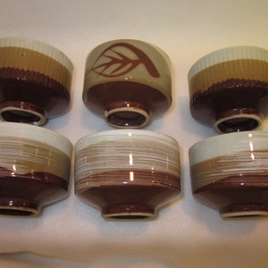 May include: Set of six brown and white ceramic teacups with different designs. The cups have a white rim and a brown base. Some cups have a leaf design, while others have a striped design.