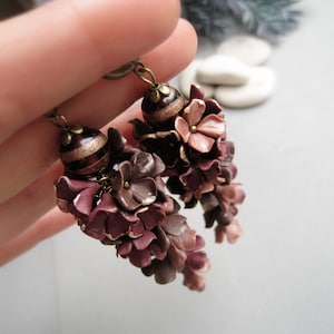 Op de afbeelding: Een paar hangende oorbellen met trossen kleine, gebeeldhouwde bloemen in tinten mauve, pruim en oudroze. Elke oorbel is bekroond met een donkerbruine bolvormige kraal, geaccentueerd door een gouden band en een messingkleurig metalen kapje.