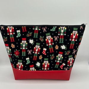 Könnte beinhalten: Schwarze Reißverschlusstasche mit rotem Boden. Die Tasche ist mit einem sich wiederholenden Muster aus Nussknackerfiguren, Zuckerstangen und Lebkuchenmännern verziert.