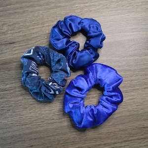 May include: Three blue hair scrunchies. One is a dark blue velvet scrunchie, one is a light blue satin scrunchie, and one is a dark blue patterned scrunchie.