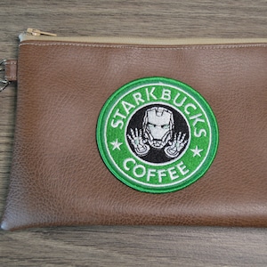 Könnte beinhalten: Braune Leder-Clutch mit goldenem Reißverschluss und Handschlaufe. Die Clutch hat einen grün-weißen gestickten Aufnäher mit den Worten "STARKBUCKS COFFEE" und einer Iron Man-Grafik.