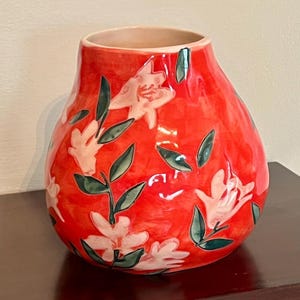 May include: A red ceramic vase with a glossy finish. The vase is adorned with white floral designs and green leaves. The vase has a rounded shape and a light beige interior.