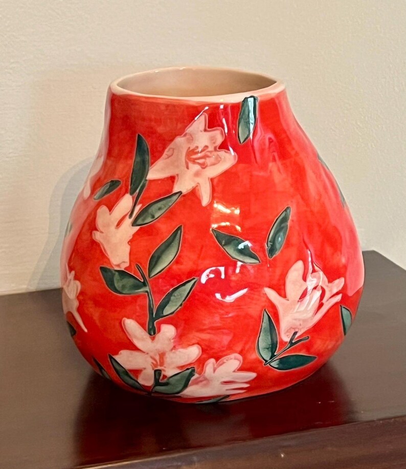 May include: A red ceramic vase with a glossy finish. The vase is adorned with white floral designs and green leaves. The vase has a rounded shape and a light beige interior.