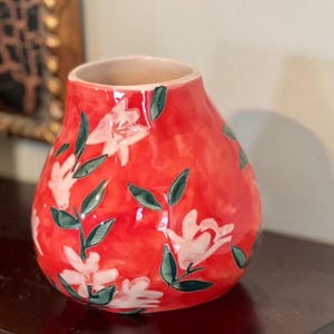 May include: A red ceramic vase with a floral design. The vase has a rounded shape with a wide base and a narrow opening. White flowers with green leaves are painted on the red surface. The vase is approximately 8 inches tall.