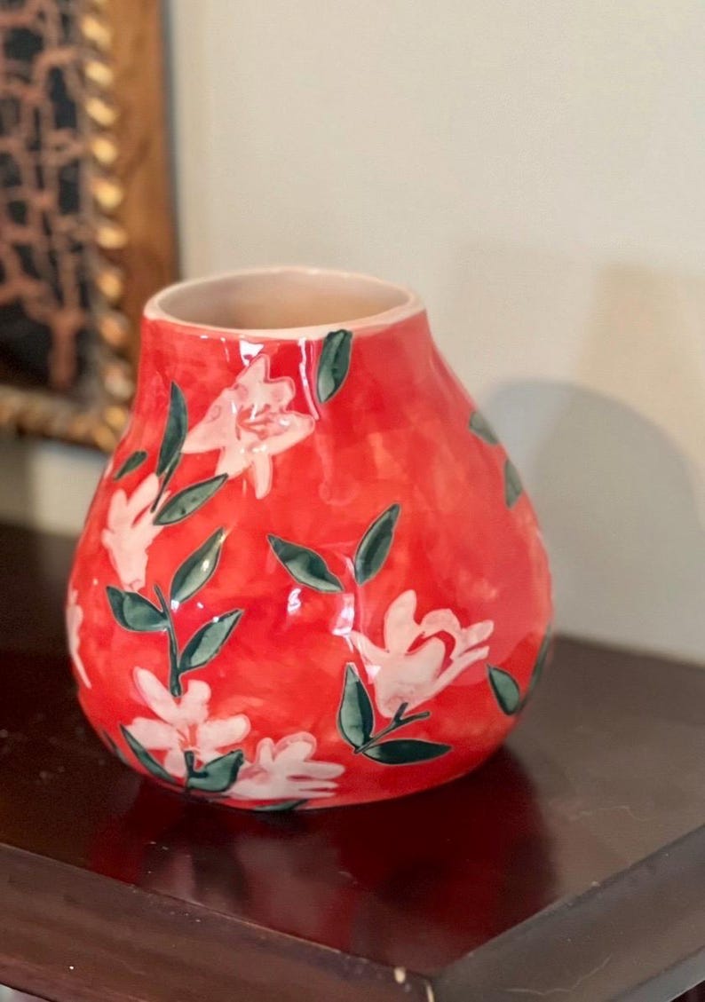 May include: A red ceramic vase with a floral design. The vase has a rounded shape with a wide base and a narrow opening. White flowers with green leaves are painted on the red surface. The vase is approximately 8 inches tall.