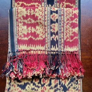 May include: A rectangular woven textile featuring a repeating geometric pattern in deep red, black, and tan. The fabric displays vertical stripes and intricate motifs, finished with a dense fringe of twisted red, black, and tan threads at the edge.