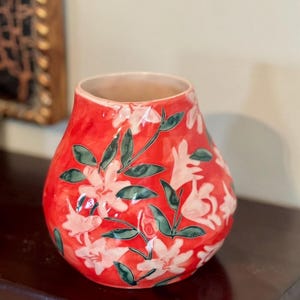 May include: A red ceramic vase with a floral design. The vase features white flowers and green leaves against a red background. The vase has a wide opening and a rounded shape, suitable for displaying flowers.
