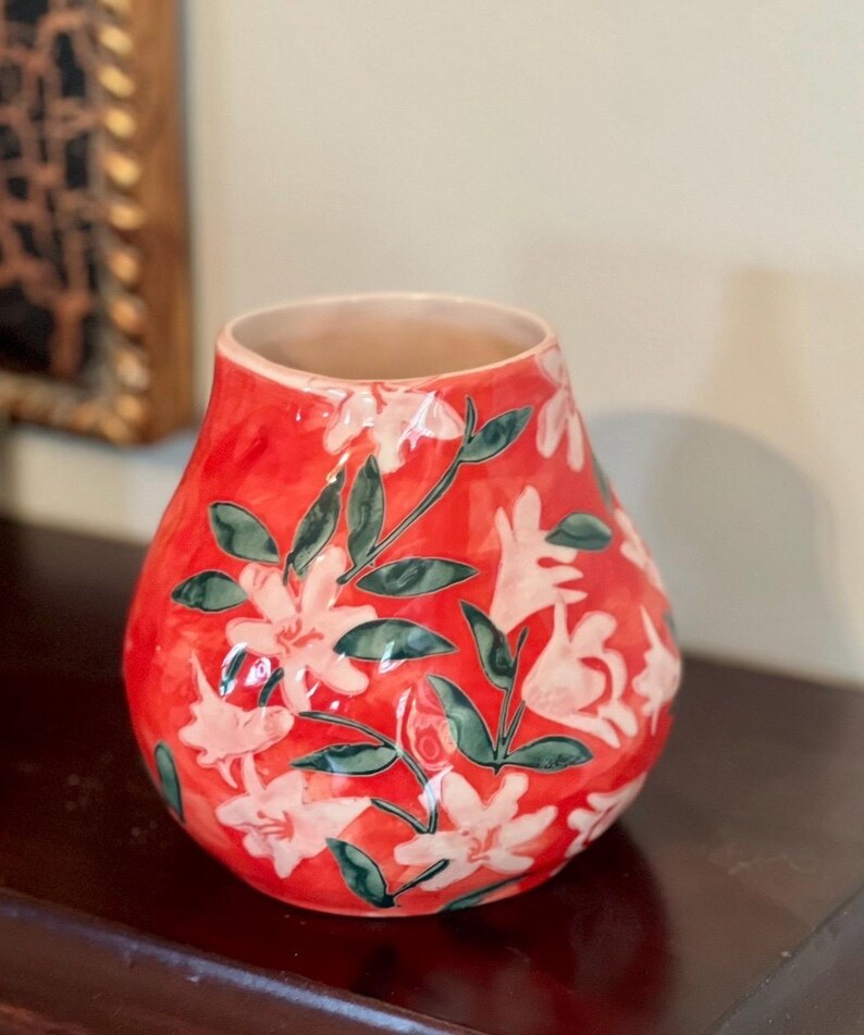 May include: A red ceramic vase with a floral design. The vase features white flowers and green leaves against a red background. The vase has a wide opening and a rounded shape, suitable for displaying flowers.