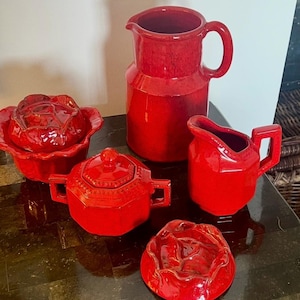May include: A collection of red ceramic kitchenware, including a pitcher, creamer, sugar bowl, and two decorative pieces. The items have a glossy finish and are arranged on a dark surface. The pieces are likely for serving or display.