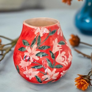 May include: A small, red ceramic vase with a floral design. The vase features white flowers and green leaves against a red background. The vase is round with a wide opening and is suitable for displaying flowers.