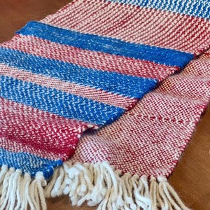May include: A woven scarf with a red, white, and blue color scheme. The scarf features a striped pattern with white fringe at the ends. The scarf is made of a soft, textured material.