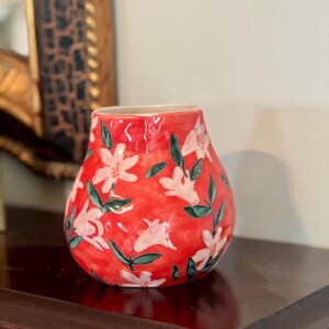 May include: A red ceramic vase with a floral pattern. The vase has a bulbous shape and is decorated with white flowers and green leaves. The vase is sitting on a dark wooden surface.