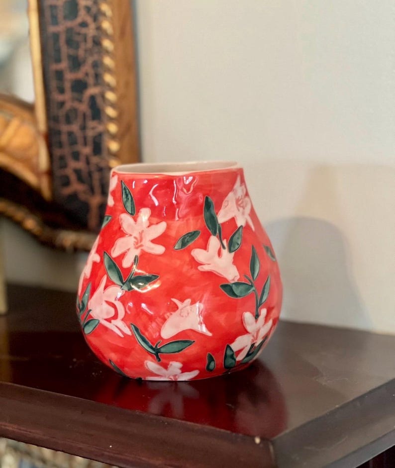 May include: A red ceramic vase with a floral pattern. The vase has a bulbous shape and is decorated with white flowers and green leaves. The vase is sitting on a dark wooden surface.