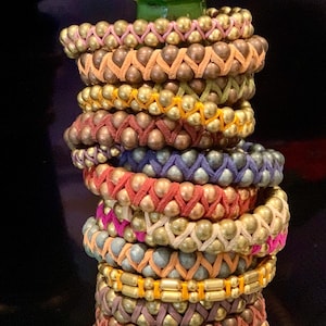 May include: A stack of beaded bracelets in various colors, including gold, orange, pink, and blue. The bracelets are made with small, round beads and woven leather or suede straps. The bracelets are stacked on a green base.