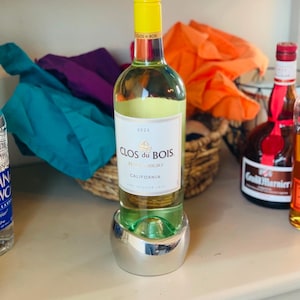 May include: A bottle of Clos du Bois Pinot Grigio wine from California, in a silver wine chiller, on a white countertop. The bottle has a yellow label with the year 2023 printed on it.