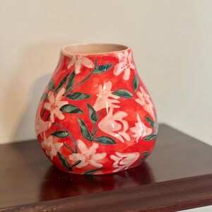 May include: A red ceramic vase with a floral pattern. The vase features white flowers and green leaves against a vibrant red background. The vase is sitting on a dark brown surface.