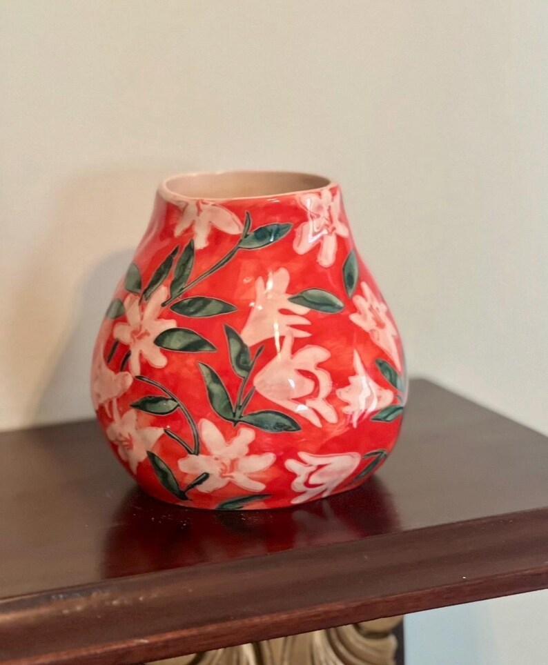 May include: A red ceramic vase with a floral pattern. The vase features white flowers and green leaves against a vibrant red background. The vase is sitting on a dark brown surface.