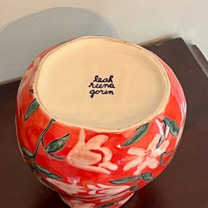 May include: A ceramic vase with a vibrant red background, adorned with white floral designs and green leaves. The interior is a light beige color. The artist's signature, "leah reena goren," is visible inside the vase.