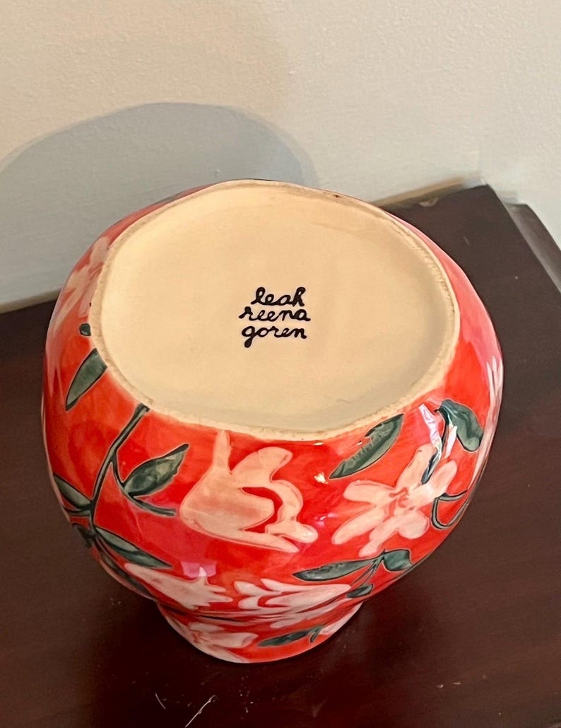 May include: A ceramic vase with a vibrant red background, adorned with white floral designs and green leaves. The interior is a light beige color. The artist's signature, "leah reena goren," is visible inside the vase.