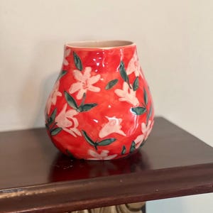 May include: A red ceramic vase with a floral pattern. The vase features white flowers with green leaves against a red background. The vase is sitting on a dark brown wooden surface.