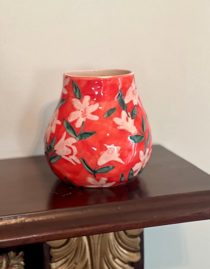 May include: A red ceramic vase with a floral pattern. The vase features white flowers with green leaves against a red background. The vase is sitting on a dark brown wooden surface.