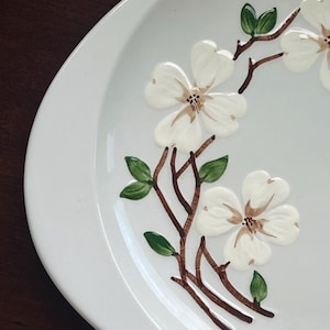 May include: White oval serving platter with a raised floral design. The design features white flowers with brown centers, brown branches, and green leaves. The platter has a smooth, glossy finish.