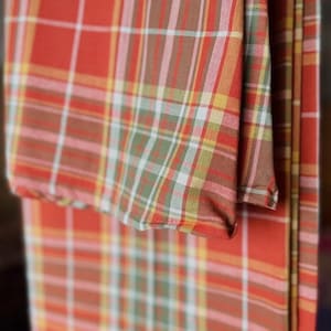 May include: A folded textile featuring a plaid pattern with intersecting lines in shades of burnt orange, muted green, yellow, and white. The fabric displays a woven texture with a consistent grid design throughout the material, showing multiple layers.