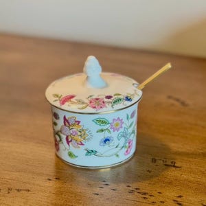 May include: A small, white ceramic sugar bowl with a lid and a gold spoon. The bowl and lid are decorated with a floral pattern in pink, yellow, and blue. The rim of the bowl and lid are trimmed with gold.