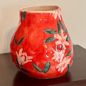 May include: A red ceramic vase with a bulbous shape and a wide opening. The vase is decorated with white floral designs and dark green leaves. The interior of the vase is a light beige color. The vase is sitting on a dark brown surface.