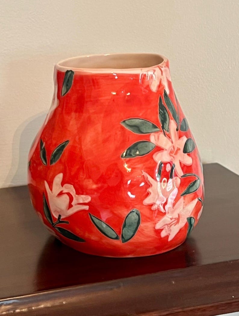 May include: A red ceramic vase with a bulbous shape and a wide opening. The vase is decorated with white floral designs and dark green leaves. The interior of the vase is a light beige color. The vase is sitting on a dark brown surface.