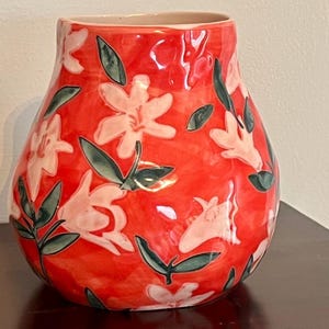 May include: A red ceramic vase with a floral design. The vase features white flowers and green leaves against a red background. The vase has a wide body and a narrow opening, suitable for displaying flowers.