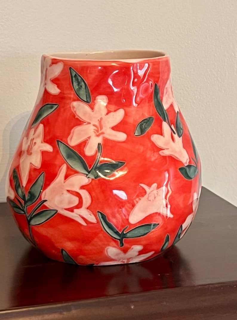 May include: A red ceramic vase with a floral design. The vase features white flowers and green leaves against a red background. The vase has a wide body and a narrow opening, suitable for displaying flowers.