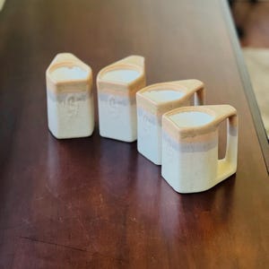 May include: Four ceramic mugs with a unique design. The mugs are off-white with a speckled texture and feature a tan rim and handle. A thin stripe of pink and gray accents the middle of the mugs. The mugs are arranged on a dark brown surface.