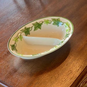 May include: Oval-shaped serving dish with a cream-colored interior and a green ivy leaf design around the rim. The dish is divided into two sections and sits on a wooden surface.