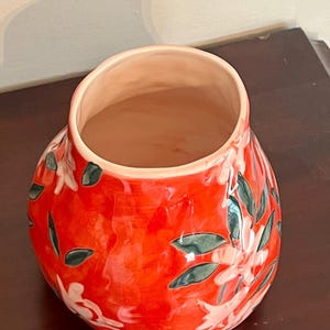 May include: A vibrant red ceramic vase with a glossy finish. The vase features a floral design with white flowers and green leaves. The interior of the vase is a light peach color. The vase is a decorative item.