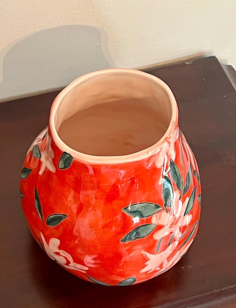 May include: A vibrant red ceramic vase with a glossy finish. The vase features a floral design with white flowers and green leaves. The interior of the vase is a light peach color. The vase is a decorative item.